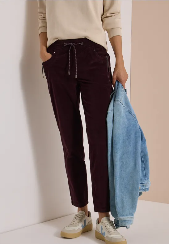 Une femme porte un pantalon velours côtelé bordeaux, un pull crème, des baskets blanches et tient une veste en jean clair.