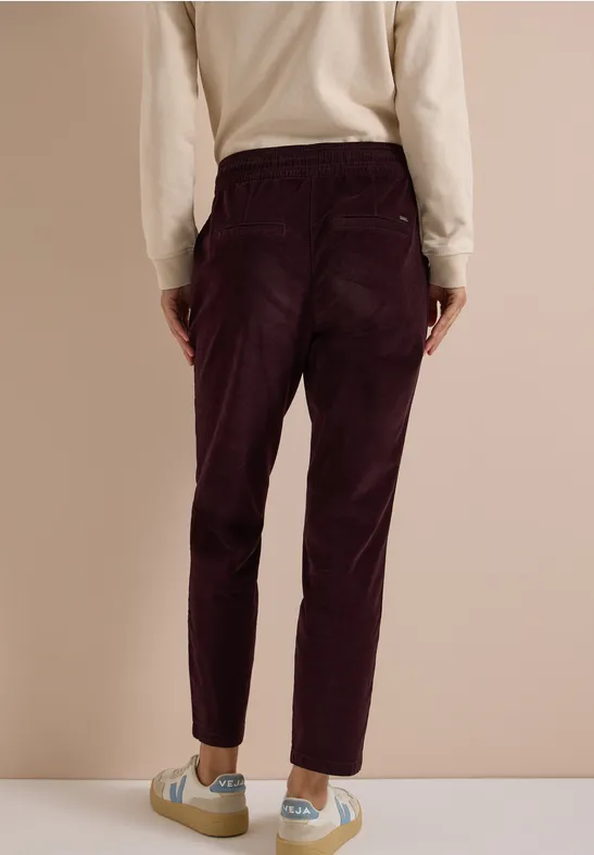 Mannequin vu de dos, portant un pantalon en velours côtelé violet foncé taille haute, un haut beige et des baskets claires.