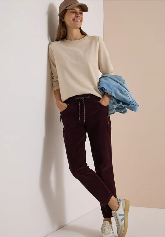 Femme appuyée contre un mur, pull beige, pantalon bordeaux, baskets et jean bleu sur le bras.