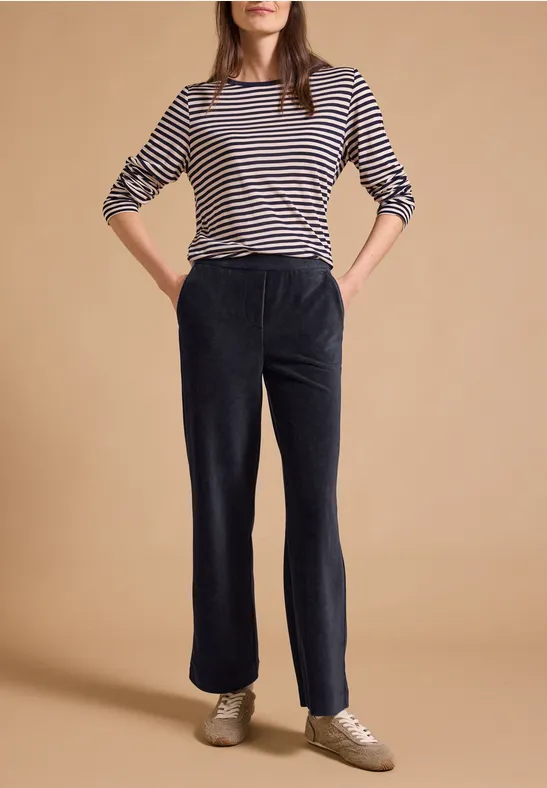 Eine Frau trägt gestreiftes 3/4-Arm-Shirt, schwarze weite Hosen und Sneakers vor hellem Hintergrund.