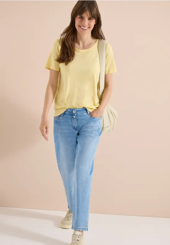 Eine Frau trägt ein gelbes T-Shirt, helle Jeans und Sneakers, mit Schultertasche vor neutralem Hintergrund.