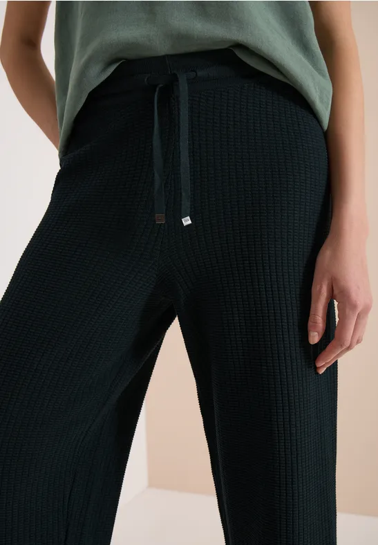 Modell trägt ärmelloses grünes Top und schwarze, gerippte Hose mit Kordelzug.