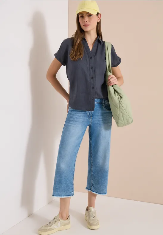 Frau lehnt an Wand, trägt schwarzes Kurzarmhemd, weite Culotte-Jeans, beige Sneaker und grüne Puffer-Tasche.