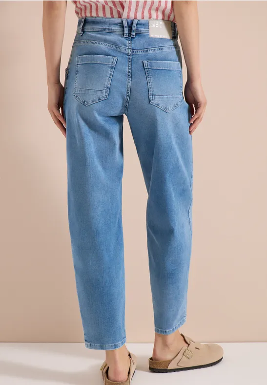 Femme de dos portant un jean taille haute bleu clair et des sandales beiges sur fond uni.