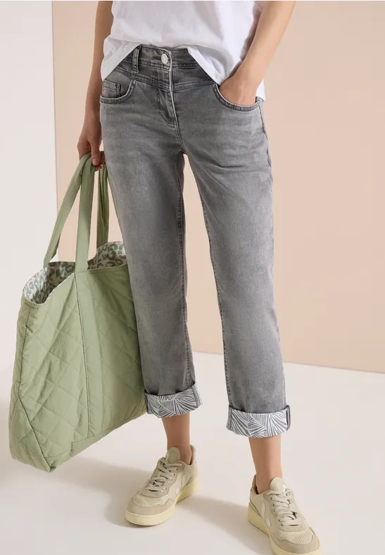 Frau trägt graue, hochgekrempelte Jeans, weißen T-Shirt, grüne Tragetasche und beige Sneaker.