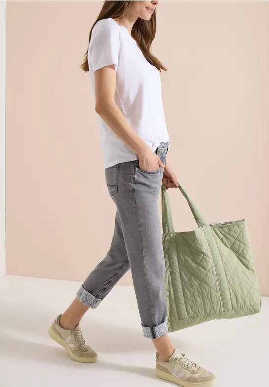 Frau in weißem T-Shirt, grauen Jeans und grünen Sneakers trägt eine große, gesteppte, grüne Tasche.