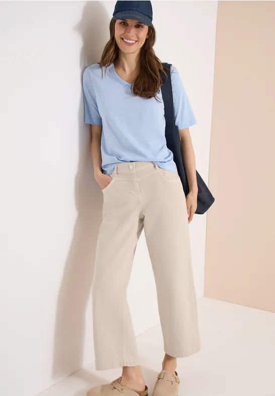 Eine Frau steht lässig an einer hellen Wand, trägt hellblaues T-Shirt, beigen Culotte-Hose, Cap und Umhängetasche.