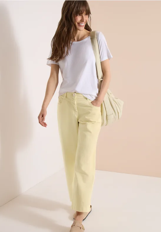 Frau trägt lässiges Outfit mit weißem T-Shirt, gelben Hosen und Umhängetasche vor hellem Hintergrund.