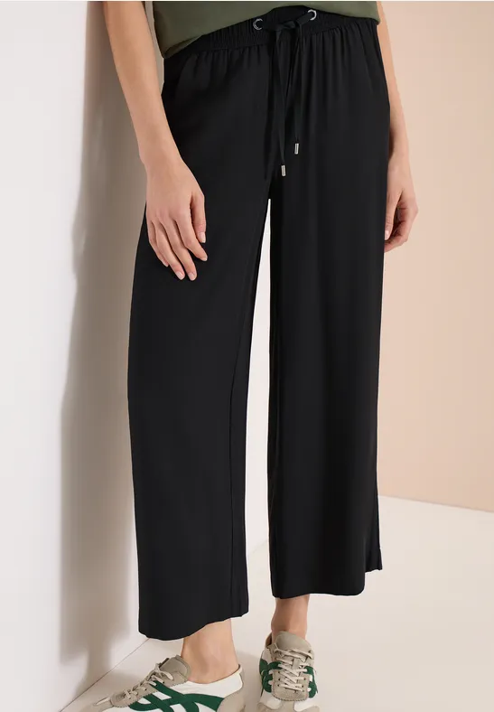 Pantalon noir ample à cordon de serrage, jambes larges, porté avec des baskets vert clair sur fond clair.