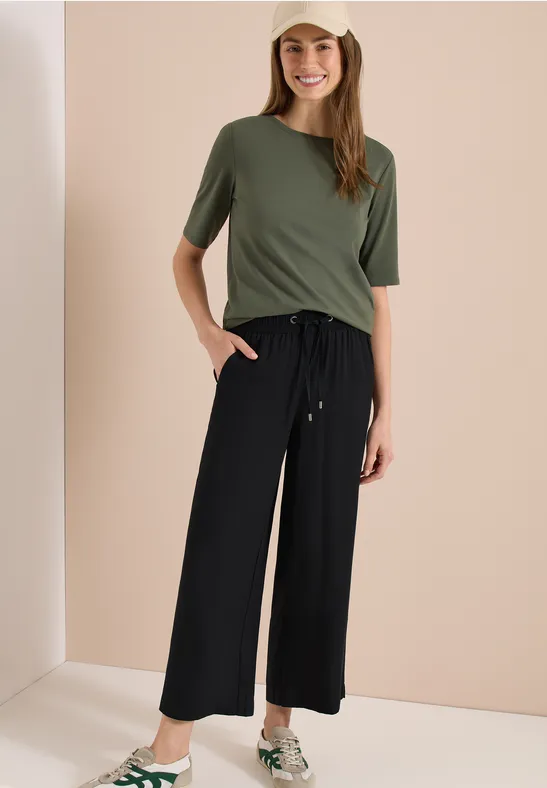 Femme portant un t-shirt olive et un pantalon large noir devant un fond neutre.