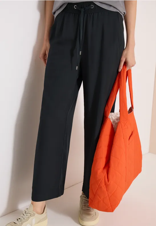 Modische schwarze weite Hose, kombiniert mit Sneakern und einer orangefarbenen Stepp-Tasche.