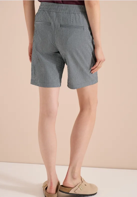 Rückenansicht einer Person in grauen, gestreiften Shorts und beigen Slippern vor hellem Hintergrund.