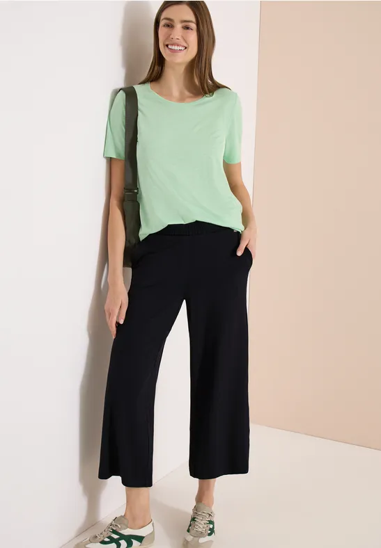 Frau lehnt an heller Wand, trägt mintgrünes T-Shirt, schwarze weite Culottes und Sandalen.