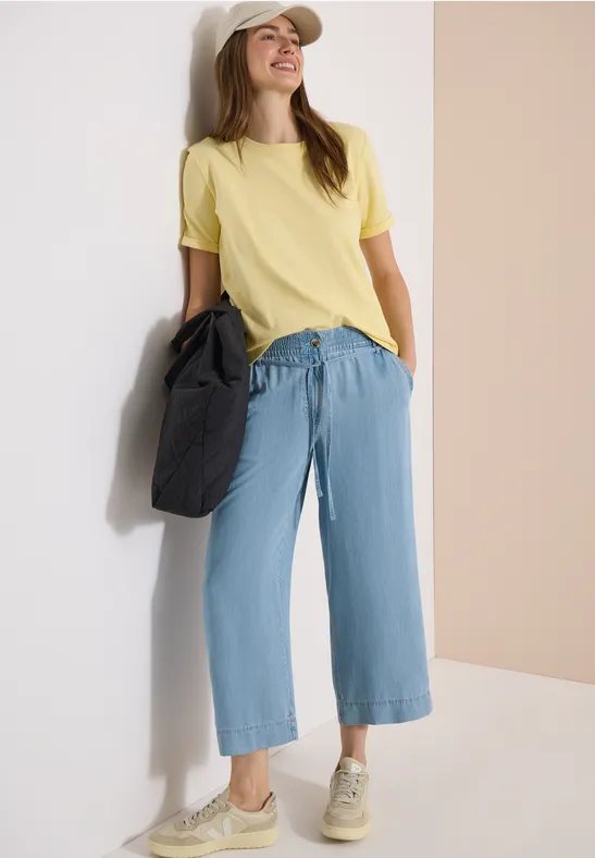 Frau lehnt an Wand mit schwarzer Tasche, gelbem T-Shirt, weiten hellblauen Jeans und Sneakers.