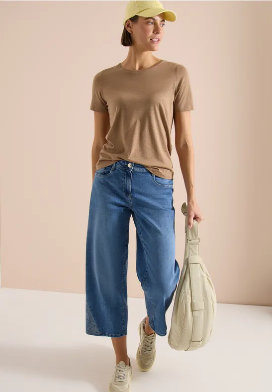 Person trägt beigefarbenes T-Shirt, weite blaue Culotte-Jeans, gelbe Kappe, trägt gepolsterte Umhängetasche und Sneaker