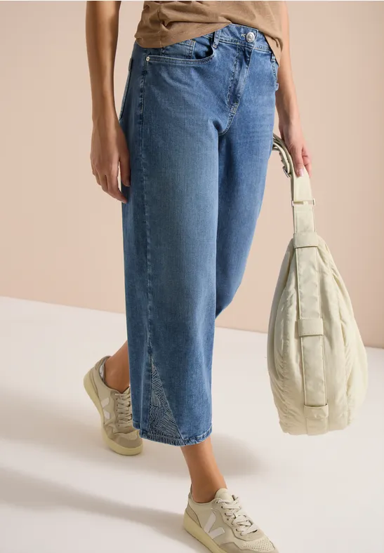 Beine in weiten Jeans, hellen Sneakern und beigefarbener, gesteppter Tasche vor neutralem Hintergrund