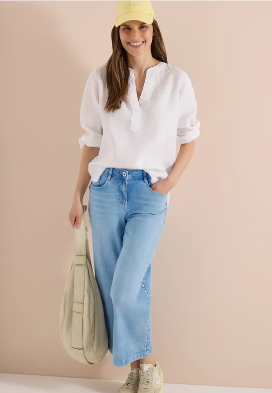 Eine Frau trägt eine weiße Bluse, helle weite Jeans und Sneaker, dazu eine grüne Tasche.