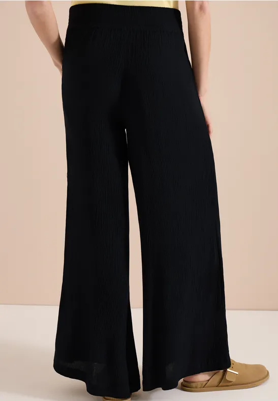 Pantalon noir ample en tricot fendu sur le côté, associé à des mocassins plats dorés sur fond neutre.