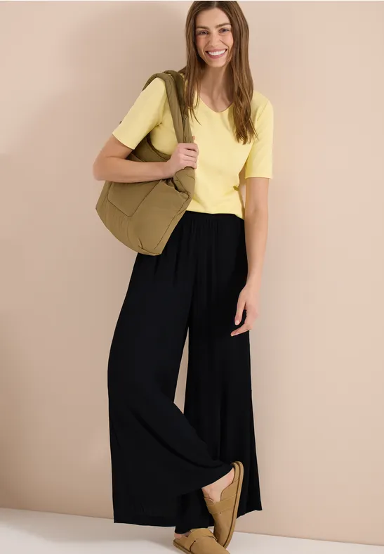 Femme en T-shirt jaune et pantalon large noir, avec un sac kaki et des espadrilles.