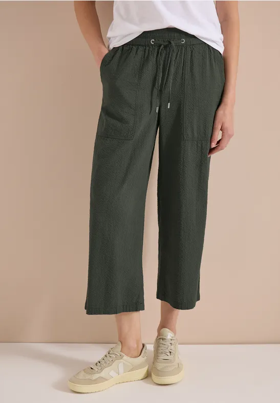 Unterschenkel einer Person in lässiger, knöchellanger, olivgrüner Hose mit Kordelzug, dazu cremefarbene Sneaker.