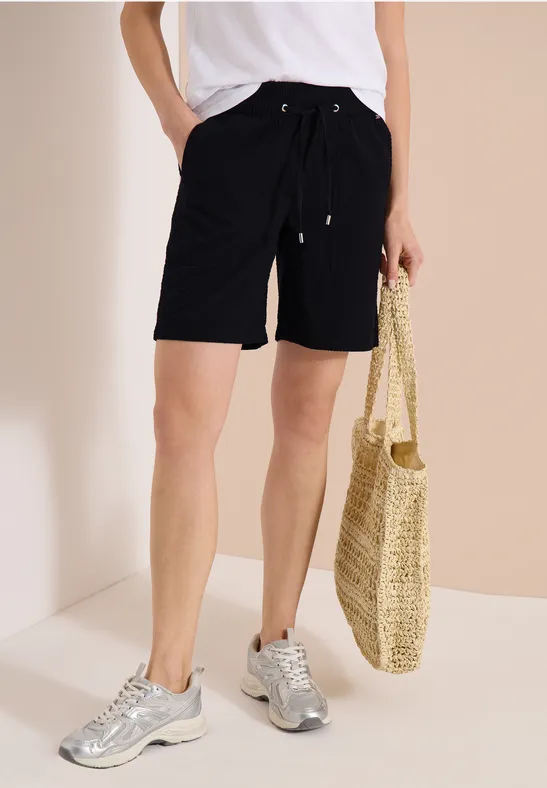 Personne en short noir et t-shirt blanc, baskets argentées, tenant un sac en paille tressée sur fond clair.