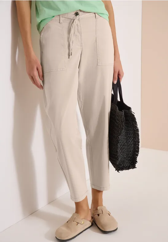 Une personne porte un pantalon beige clair confortable avec des poches, des chaussures beiges et tient un sac noir.