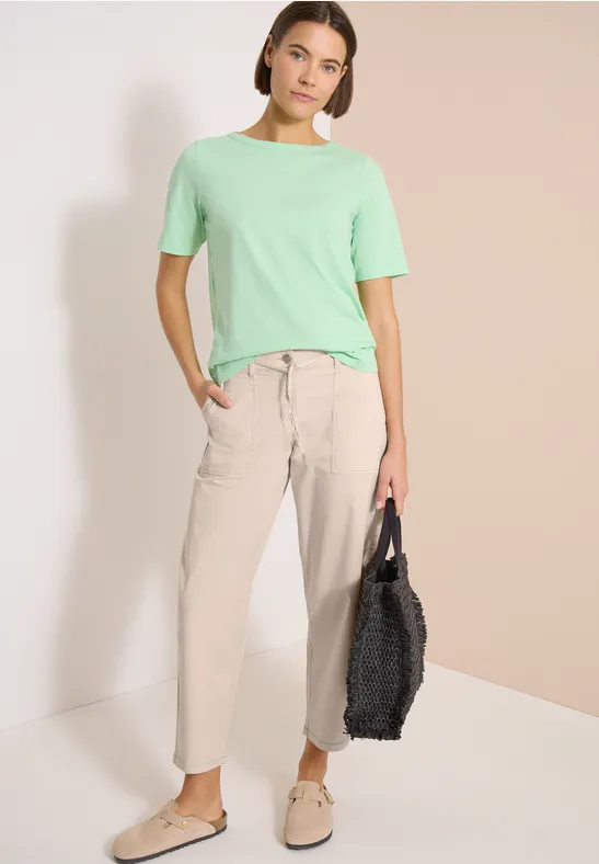 Personne devant fond clair, porte t-shirt menthe, pantalon beige et chaussures, tient sac noir.