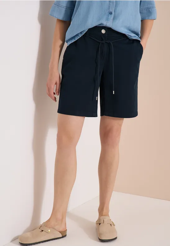 Mann trägt hellblaues Kurzarmhemd, dunkelblaue Shorts mit Kordelzug und beige Slipper vor neutralem Hintergrund.