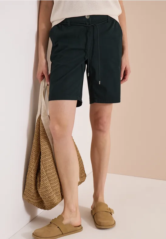 Dame in schwarzen Shorts, weißem Top, trägt Strohtasche und Slipper vor neutralem Hintergrund