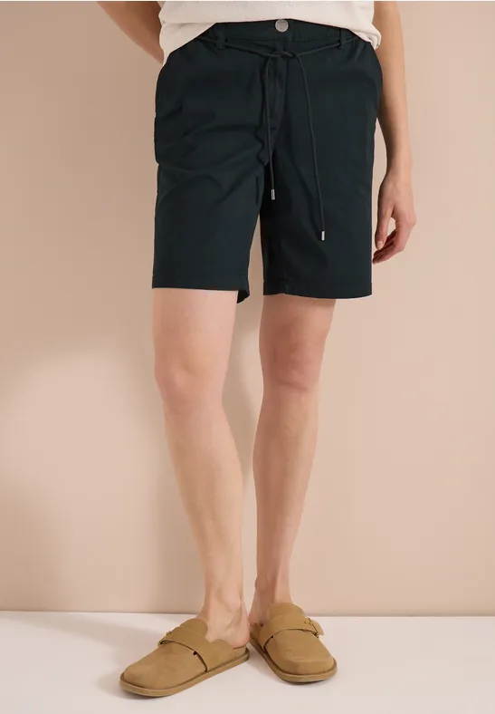 Person trägt schwarze, knielange Shorts mit Tunnelzug und beige Slipper vor neutralem Hintergrund.