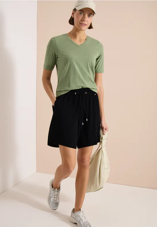 Personne portant un t-shirt vert en V, short noir, baskets argentées et un sac en toile sur fond neutre.