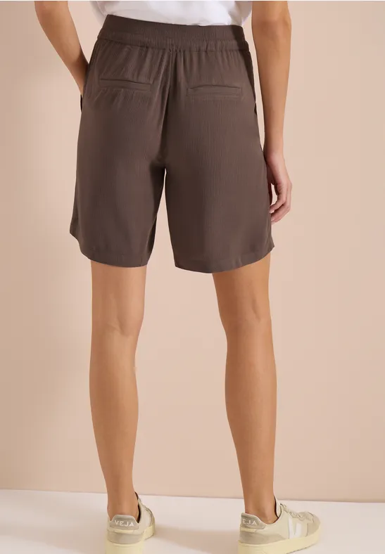 Rückenansicht einer Person in lässigen braunen Shorts mit elastischem Bund und Taschen vor beigem Hintergrund.