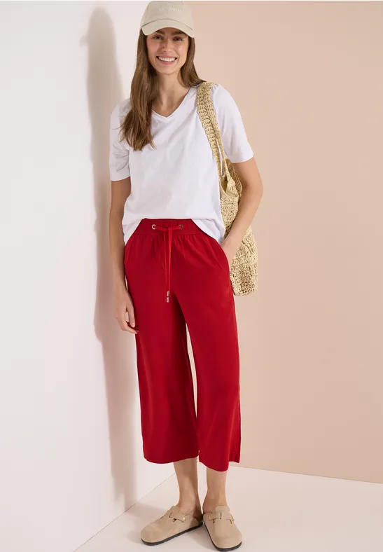 Frau lehnt an Wand, trägt weißes T-Shirt, rote Caprihose, Strohtasche, Espadrilles und Strohhut.