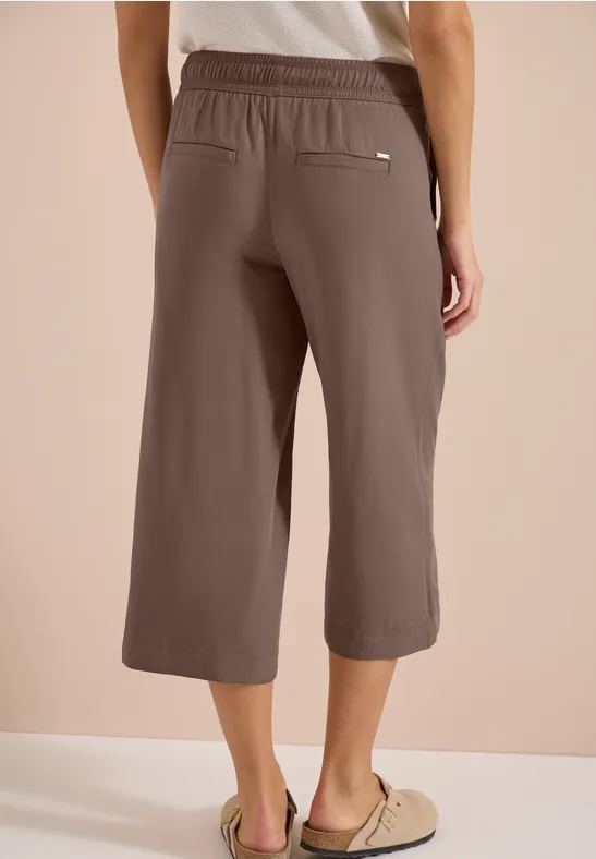 Frau von hinten in braunen, knöchellangen Culottes mit elastischem Bund, Eingrifftaschen und Sandalen.