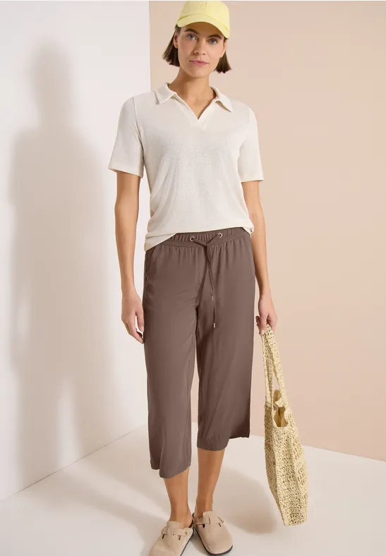 Frau trägt weißes Poloshirt und braune 7/8-Hose, hält geflochtene Einkaufstasche, lässiger Look.