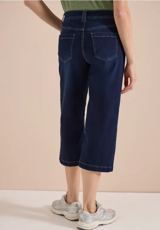 Rückenansicht einer Person in knielangen Jeanshosen und Sportschuhen vor neutralem Hintergrund.