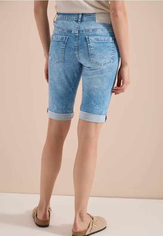 Frau von hinten in hellblauen, knielangen Jeans-Shorts mit Umschlag und beigen Loafern vor hellem Hintergrund.