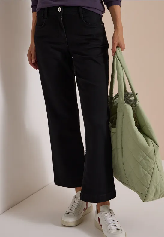Vue des jambes en jean noir et baskets blanches, avec un sac matelassé vert.