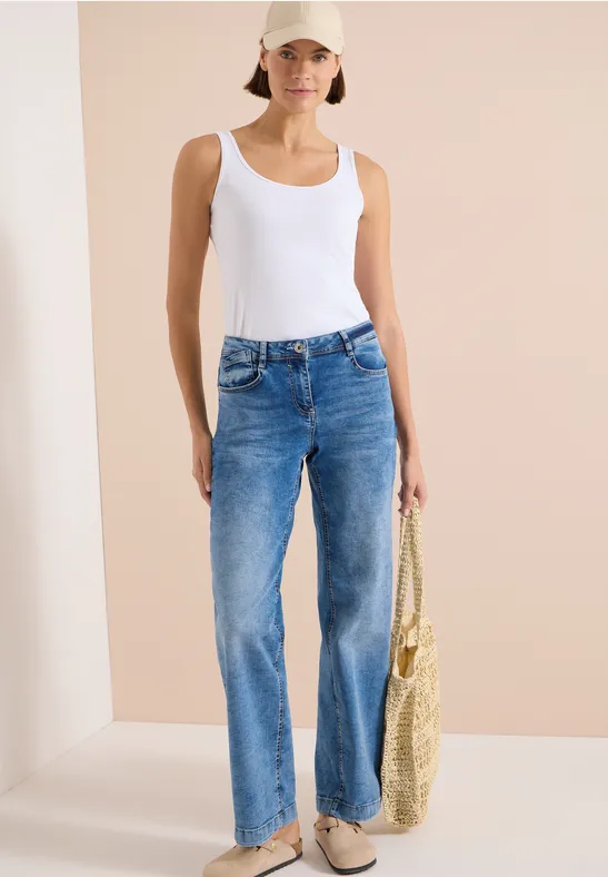 Frau im weißen Top und blauen Jeans, die eine Strohtasche hält.