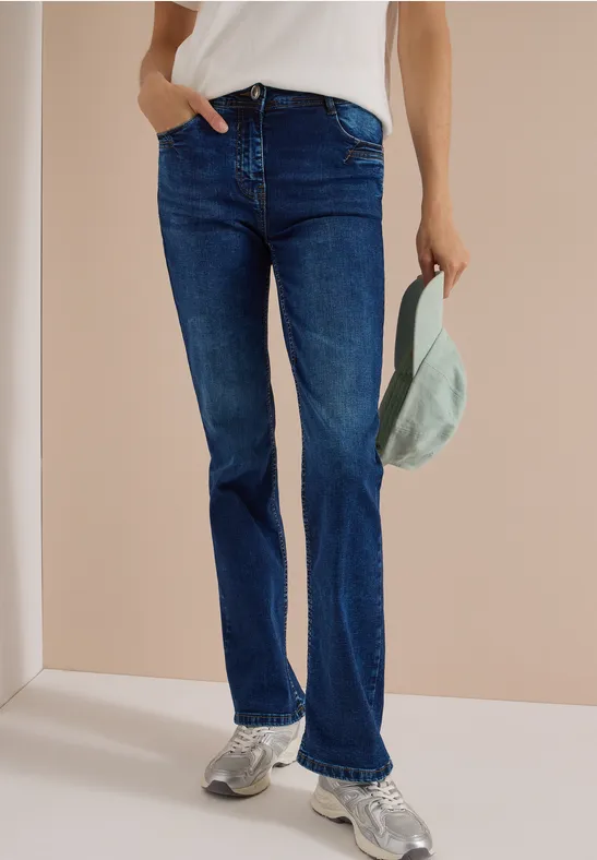 Person trägt blaue Jeans, weißen Oberteil und Sneaker, hält eine Schirmmütze in der Hand.