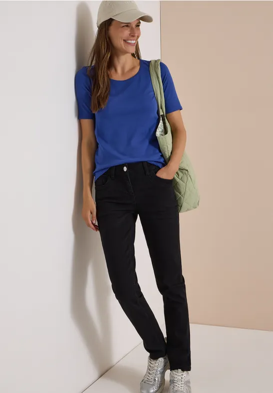 Eine Frau lehnt lächelnd an einer Wand, trägt ein helles Cap, blaues Shirt, schwarze Hose und grüne Tasche.