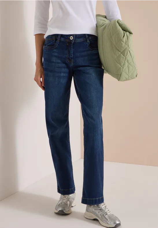 Personne porte un jean droit foncé, un haut blanc, des baskets argentées et tient un sac matelassé vert.