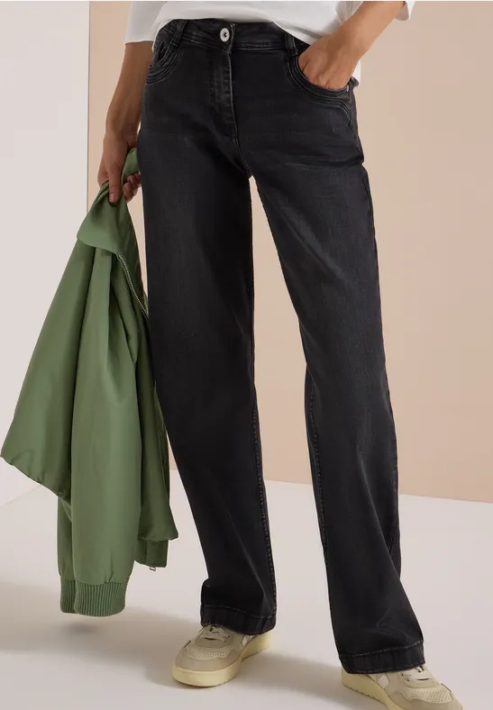 Personne porte un jean noir droit, tient une veste verte à la main et des baskets claires aux pieds.