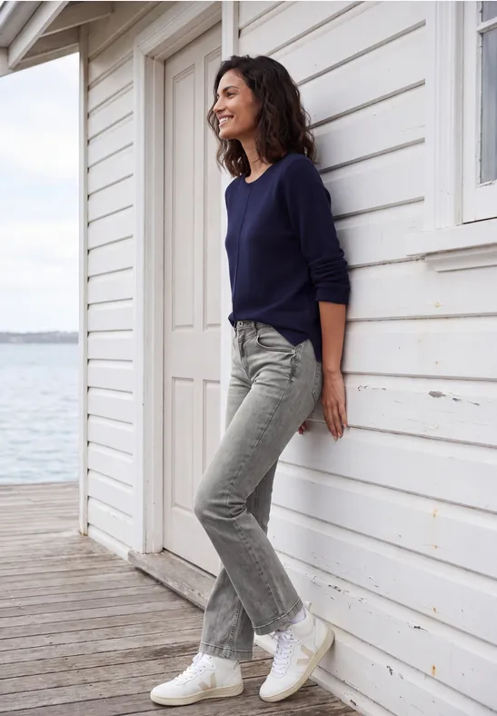 Femme en jean et pull, souriante, appuyée contre une blanche façade en bois au bord de l'eau.