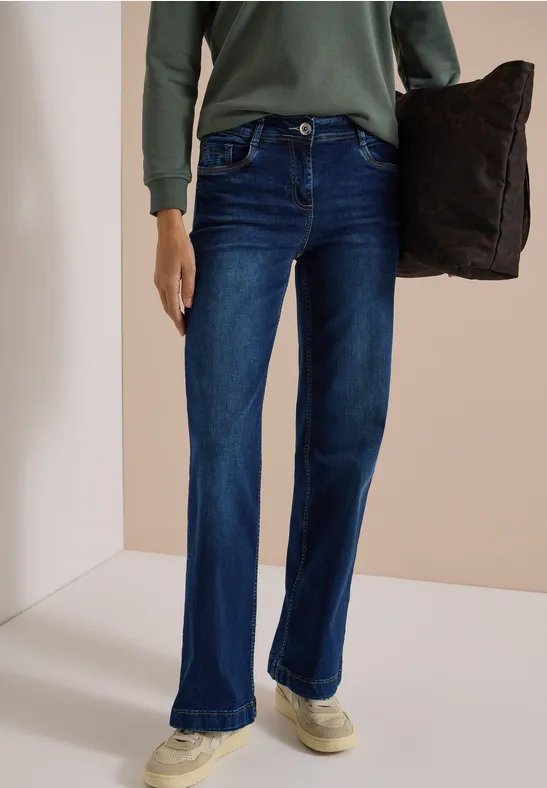 Personne porte un jean évasé bleu foncé, un haut vert à manches longues et tient un coussin foncé.