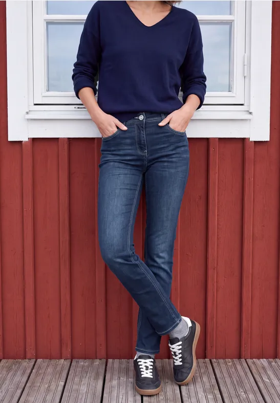 Personne en pull bleu foncé, jean et baskets, appuyée sur une façade en bois rouge avec une fenêtre.
