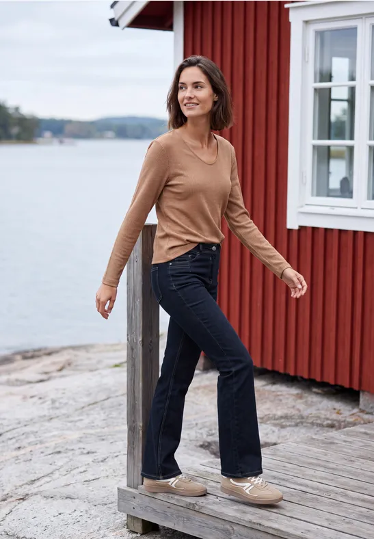 Femme en pull marron et jean foncé souriante, marchant sur un ponton en bois devant une cabane rouge au bord de l’eau.