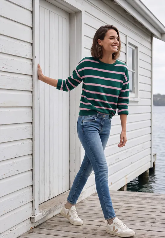 Femme en jean et pull rayé marchant en souriant devant une cabine blanche au bord de l’eau.