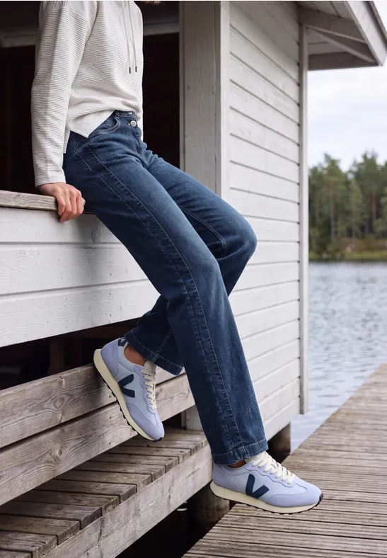 Person in Jeans und weißen Sneakers sitzt entspannt auf einem Holzsteg am ruhigen Seeufer.
