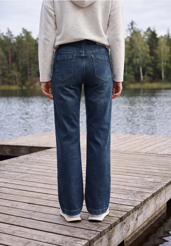 Eine Person steht von hinten auf einem Holzsteg am Seeufer und trägt blaue Jeans und einen hellen Kapuzenpullover.